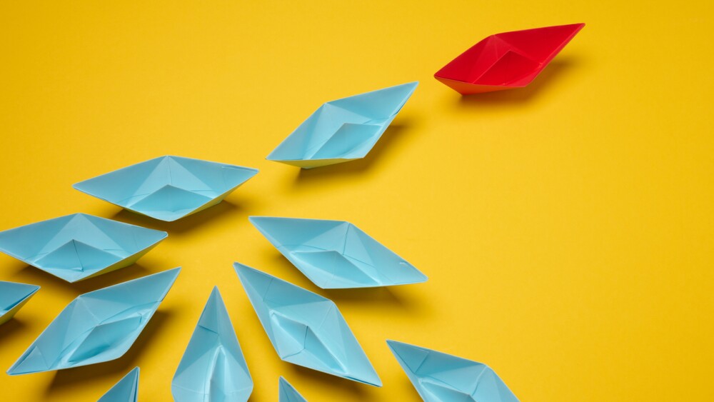 Group of blue paper boats following on red on yellow background, mass manipulation
