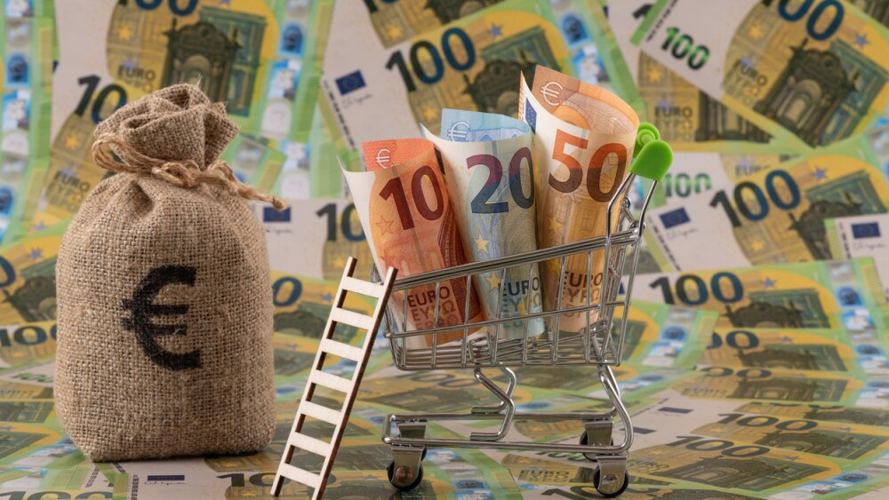 A conceptual story about finance in the Eurozone, featuring €10, €20, and €50 notes in a supermarket shopping cart, with €100 notes in the background.