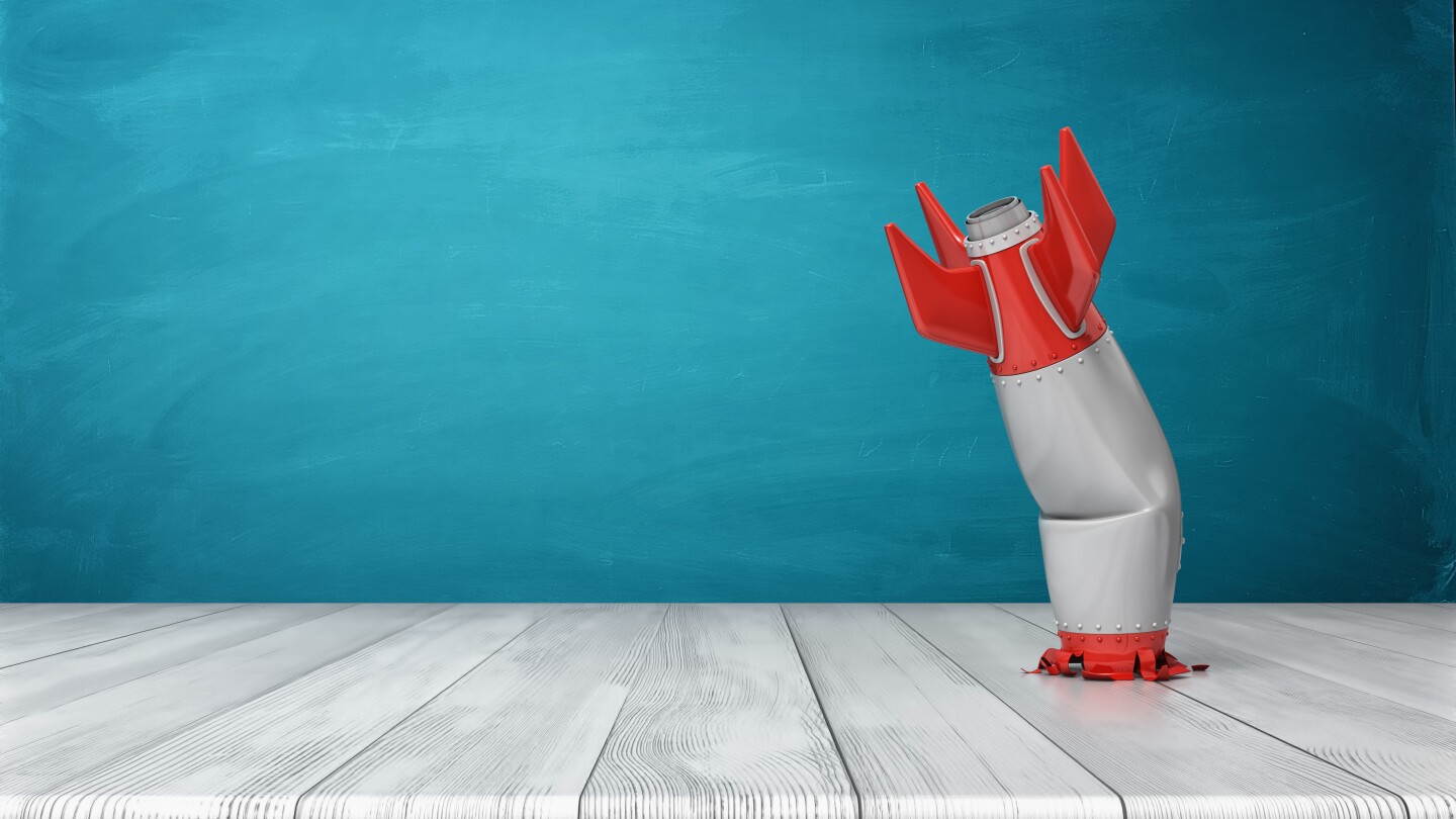 3d rendering of a red and silver realistic model of a retro rocket stands crashed into a wooden desk on a blue background. Failed launch. Technological progress. Technical mistake.