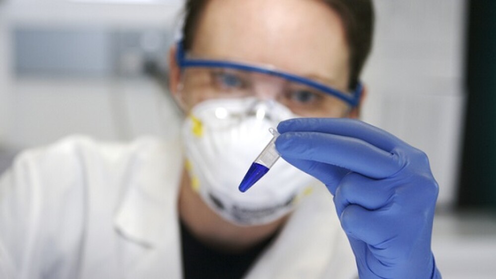 "Scientist with a labcoat, gloves, face mask, and goggles examines a small tube filled halfway with blue liquid."