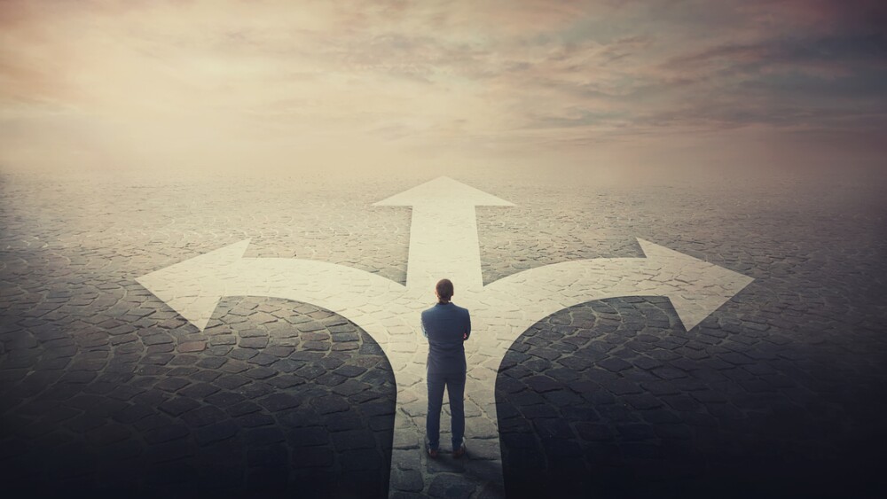 Doubtful businessman stands in front of a crossroad with road split in three different ways as arrows shows opposed courses. Business challenge, choosing a correct pathway. Difficult decision concept