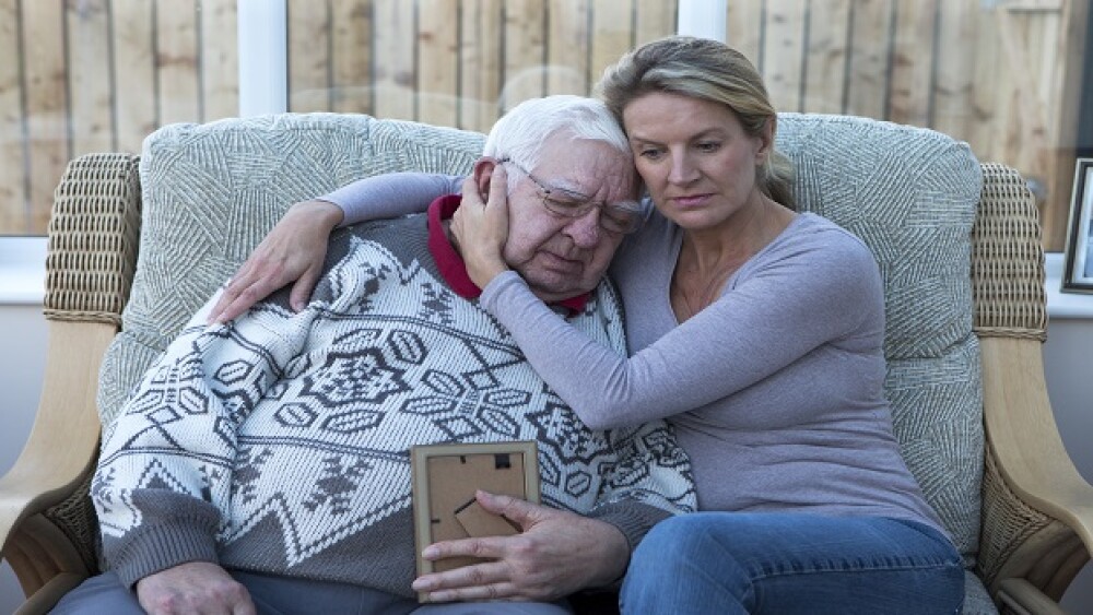 A shot of a senior man sitting on the sofa with his daughter, looking at a photograph. He is cuddling his daughter, looking sad and holding the photo.