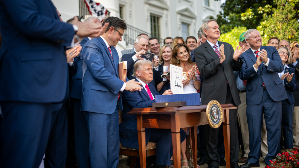 Donald Trump sits at a desk holding up a piece of paper he just signed while a crowd of people applauds