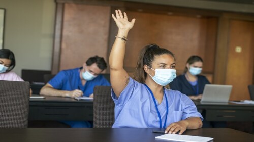 A female medical student attending a school lecture raises her hand to ask a question. The multi-ethnic group of adult students is wearing medical scrubs and protective face masks to prevent viral infection during the Covid-19 pandemic.
