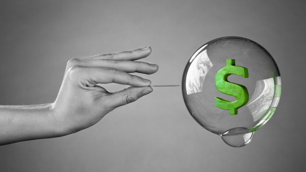 A hand about to burst a money bubble with a pin. Metaphor for an economic or financial bubble crisis.