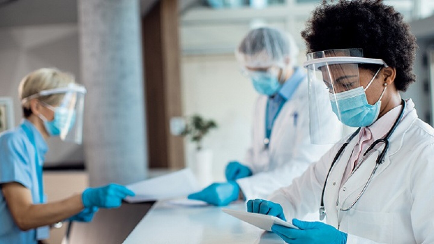 Black female doctor using digital tablet while standing at reception desk at medical clinic and wearing face shield and mask due to COVID-19 pandemic.