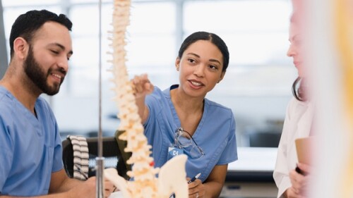 The young adult female medical student asks the mid adult female teacher a question about the human spine as her young adult male friend listens.