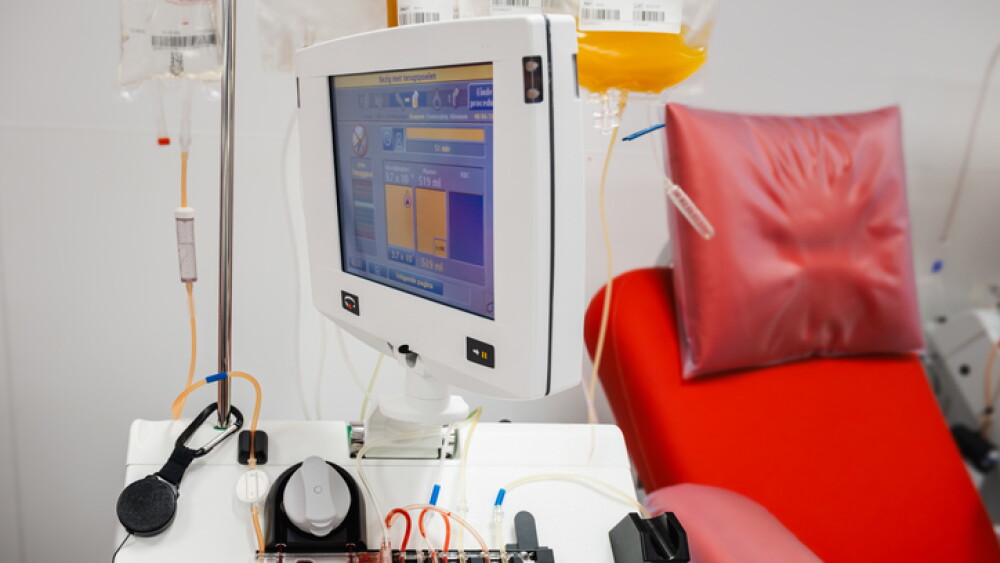 An apheresis machine with a screen and plasma bags on an IV stand beside a red donation chair at a blood bank, illustrating automated plasma collection equipment.