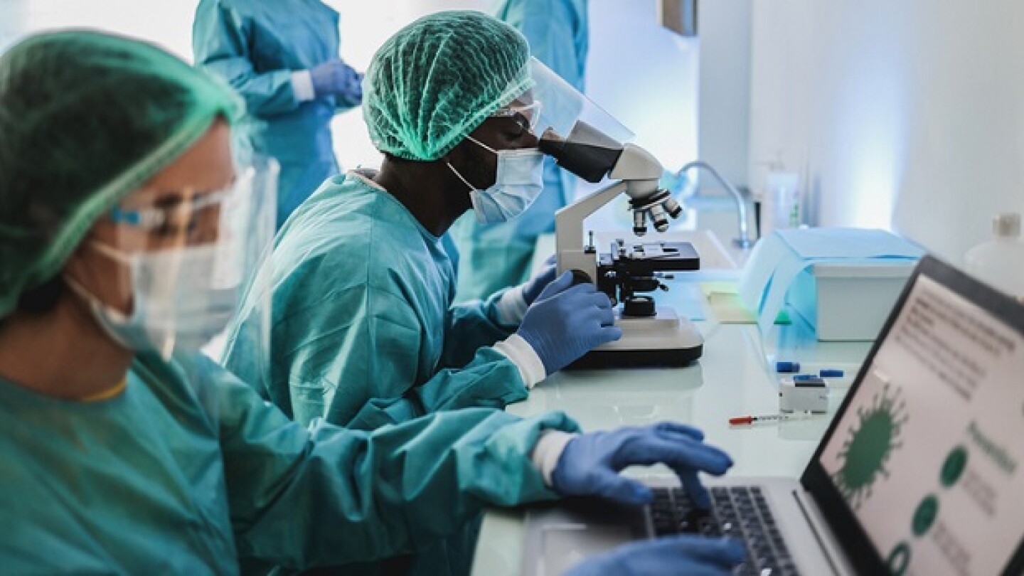 Multiracial medical scientists in hazmat suit working with microscope and laptop computer inside modern laboratory hospital - Focus african man face