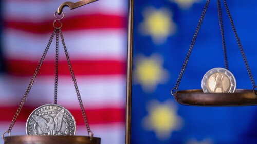 US Dollar coin and EU currencies on weighing scale against Unated States and European Union flags as symbol of trade war, economic conflict. Selective focus on Dollar.