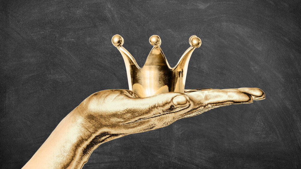 Gilded female hand holding a golden crown with dark chalkboard background. Royalty, succes and high quality concept.