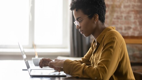 Side view focused young African American businesswoman in eyewear working on computer, sitting at table in modern loft office room, typing message, preparing electronic document or presentation.