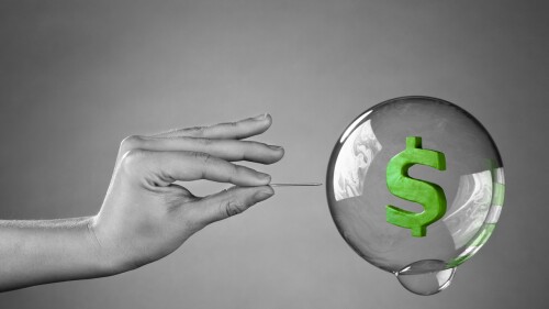 A hand about to burst a money bubble with a pin. Metaphor for an economic or financial bubble crisis.
