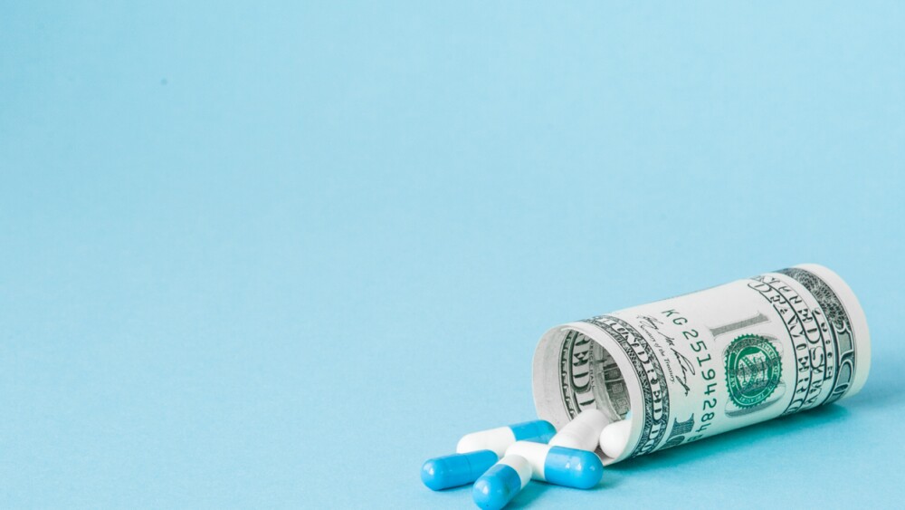Money dollar rolled up with pills flowing out isolated on blue background, high costs of expensive medication concept. Copy space.