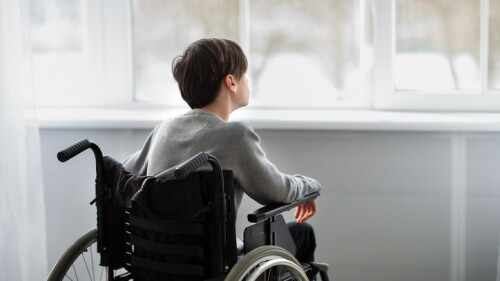 Back view of disabled teenager in wheelchair looking out window, having depression at home, copy space. Paraplegic teen boy feeling stressed and upset, suffering from loneliness indoors