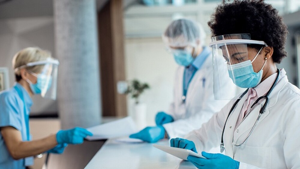 Black female doctor using digital tablet while standing at reception desk at medical clinic and wearing face shield and mask due to COVID-19 pandemic.