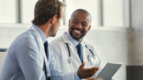 Two mature smiling doctors having discussion about patient diagnosis, holding digital tablet. Successful african surgeon discussing case after positive result. Representative pharmaceutical discussing with african happy doctor about new medicine.