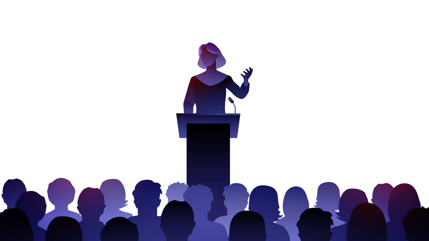 Female Public Speaker On Lectern In Front Of Audience.
