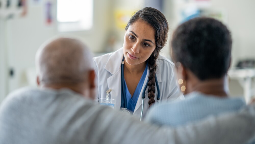 A female doctor of mixed race, meets with a senior couple as she reviews recent test results with them. The senior couple of African decent, listens closely as they receive the good news.