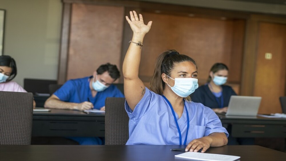 A female medical student attending a school lecture raises her hand to ask a question. The multi-ethnic group of adult students is wearing medical scrubs and protective face masks to prevent viral infection during the Covid-19 pandemic.