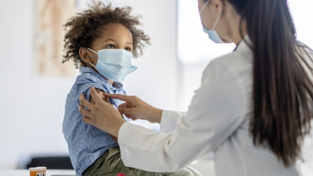 A sweet little mixed race boy sits up on an examination table as he receives an immunization. He is looking up and smiling as the doctor places the bandage on his arm. Both are wearing medical masks to protect them from COVID.