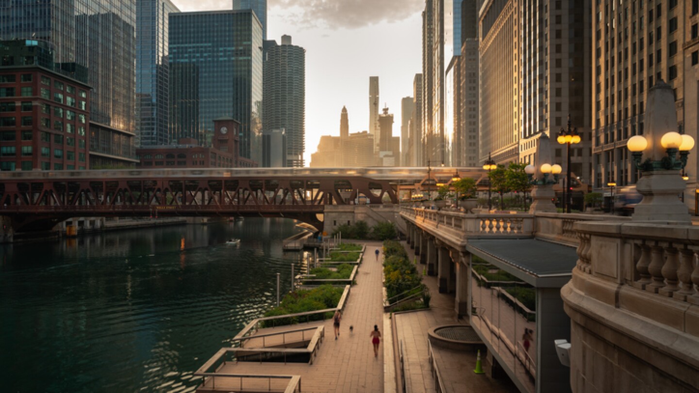 Beautiful downtown Chicago morning along the river as people jog on the path below and train crosses a bridge as the sun casts yellow light into the scene from behind the high-rise buildings beyond.
