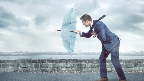 A young business man holding an umbrella to shield himself against a strong headwind. Conceptual image depicting adversity.