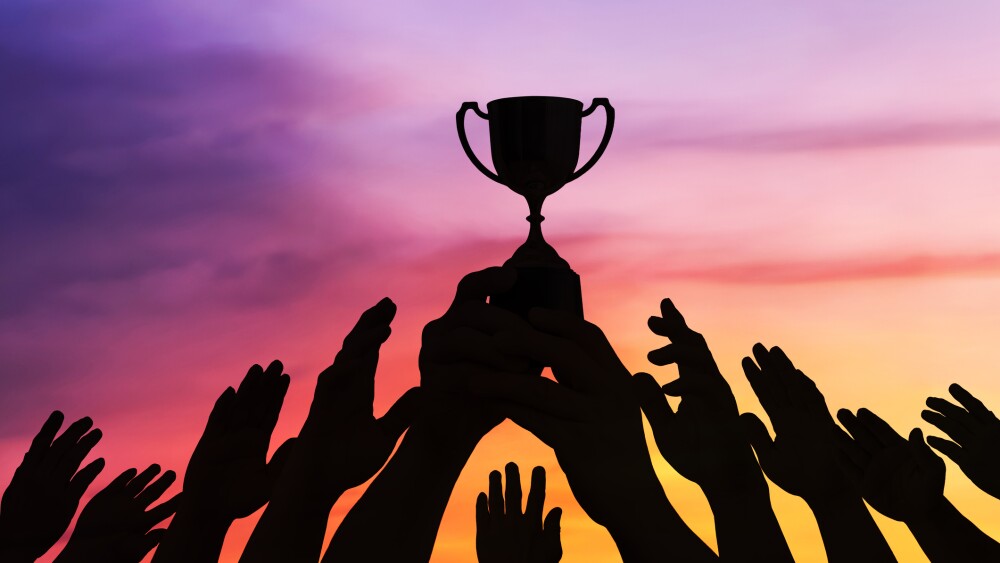 Crowd silhouettes reach toward a raised trophy beneath a vivid sunset sky, showing shared ambition, victory, teamwork, celebration, and success with open space for text.