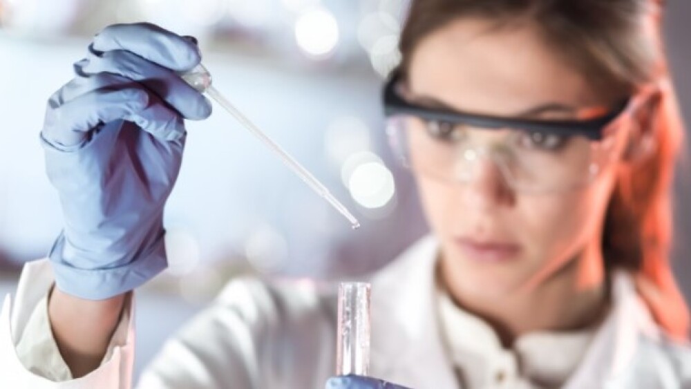Pictured: Woman in lab coat, safety glasses, and g