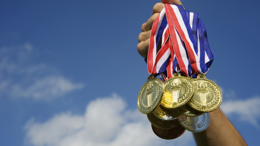 Hand holds up a bunch of gold, silver, and bronze medals in bright blue sky